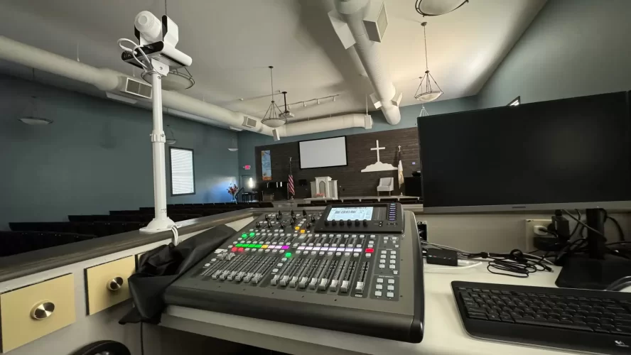 A church live stream setup featuring a digital audio mixing console on a desk, a computer monitor, a keyboard, and a PTZ auto-tracking camera mounted on a pole. The background shows a church interior with pews, a pulpit, a cross on the wall, and a projection screen.