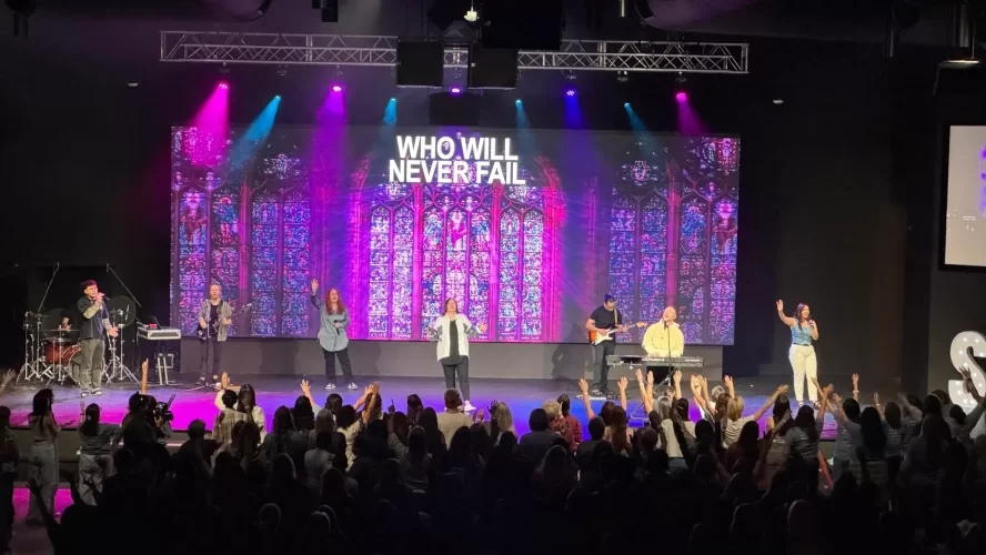 Contemporary church sanctuary, interior, several worship singers performing in front of a large LED video wall with a purple stained glass design, with church audio visual equipment such as LED stage lights and subwoofer speakers hung above them