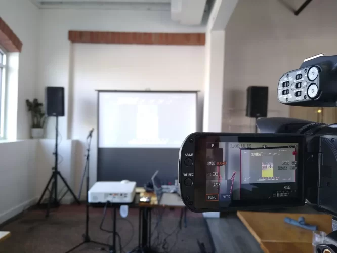 A hybrid event production setup in a small venue, featuring a camcorder recording a projection screen and stage area with speakers, microphones, and AV equipment.